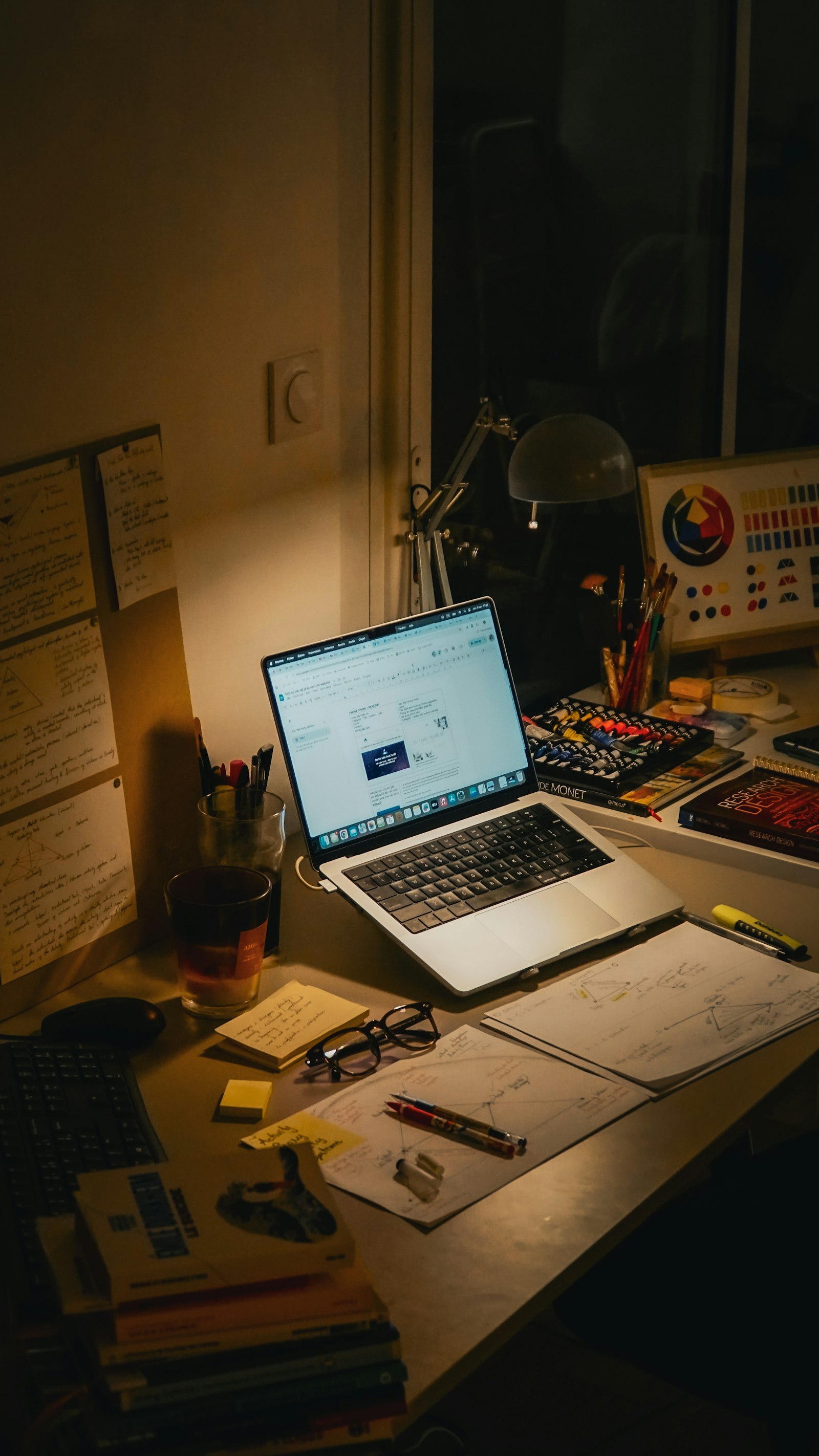 A desk with a laptop, books and papers, lit at night